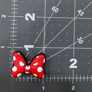 Puede incluir: Un dije de zapato en forma de lazo rojo con lunares blancos y borde negro. El dije mide aproximadamente 5 cm de ancho y est&aacute; sobre un fondo de cuadr&iacute;cula negra.