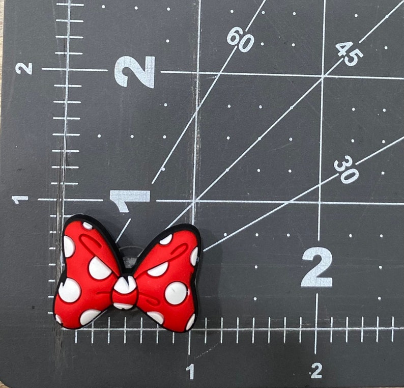 Puede incluir: Un dije de zapato en forma de lazo rojo con lunares blancos y borde negro. El dije mide aproximadamente 5 cm de ancho y est&aacute; sobre un fondo de cuadr&iacute;cula negra.