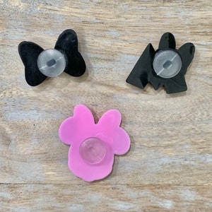 Puede incluir: Se muestran tres dijes para zapatos. Uno es un lazo negro, otro una forma geom&eacute;trica negra y el tercero un lazo rosa. Cada dije tiene un accesorio redondo y transparente en la parte posterior.