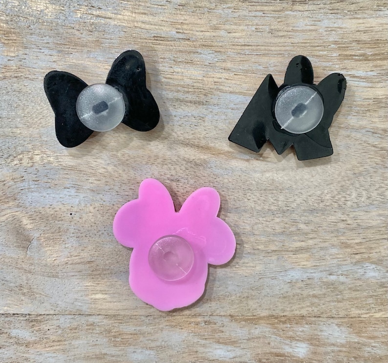 Puede incluir: Se muestran tres dijes para zapatos. Uno es un lazo negro, otro una forma geom&eacute;trica negra y el tercero un lazo rosa. Cada dije tiene un accesorio redondo y transparente en la parte posterior.