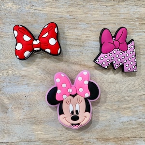 Puede incluir: Tres dijes de zapatos coloridos con dise&ntilde;os ic&oacute;nicos. Un lazo rojo con lunares blancos, un lazo rosa en una letra "M" con lunares blancos y una cara de Minnie Mouse con un lazo rosa y lunares blancos.