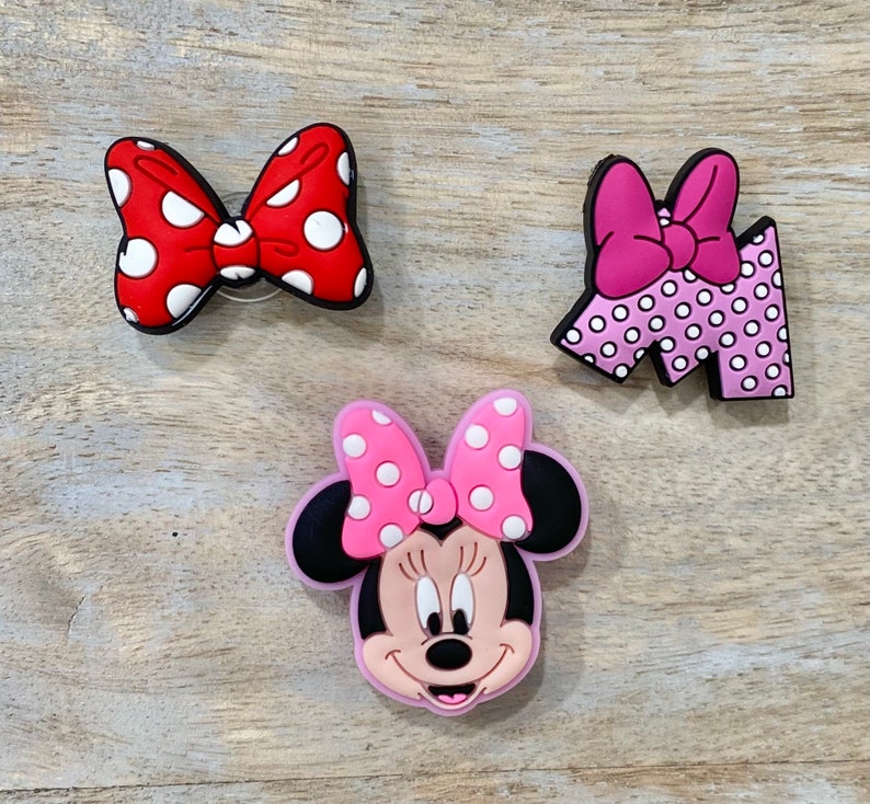 Puede incluir: Tres dijes de zapatos coloridos con dise&ntilde;os ic&oacute;nicos. Un lazo rojo con lunares blancos, un lazo rosa en una letra "M" con lunares blancos y una cara de Minnie Mouse con un lazo rosa y lunares blancos.