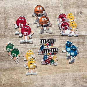 May include: A collection of colorful M&M's character charms in various colors, including red, orange, yellow, green, and blue. Some charms feature the M&M's World logo. The charms are on a light brown wooden surface.