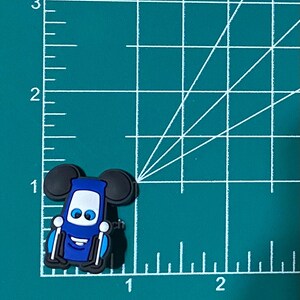May include: Blue and black cartoon character charm with Mickey Mouse ears. The character has a smiling face with white eyes and a blue body. The charm is made of rubber.