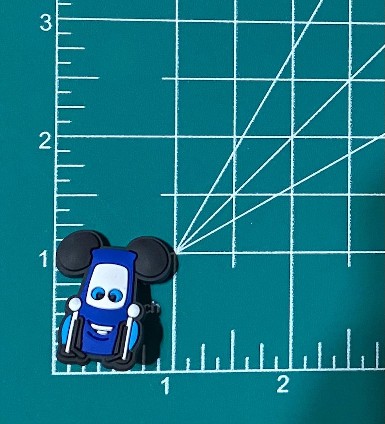 May include: Blue and black cartoon character charm with Mickey Mouse ears. The character has a smiling face with white eyes and a blue body. The charm is made of rubber.