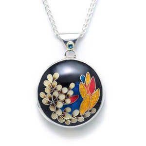 Puede incluir: Un collar de cadena de plata con un colgante redondo con fondo negro y un diseño floral colorido. El colgante tiene una piedra preciosa azul en la parte superior.