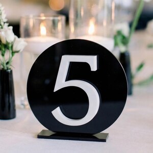May include: A black and white table number 5, displayed on a white tablecloth. The number is in a white sans-serif font, set against a black circular background. Small black vases with white flowers are in the background.