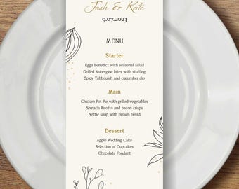 May include: A wedding menu card on a white plate with a fork and knife on a wooden table. The menu card has the names "Josh & Kate" and the date "9.07.2023" in gold script. The menu lists starters, main courses, and desserts.