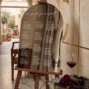 Könnte beinhalten: Ein verspiegelter Sitzplan mit Gästenamen auf einer Holzstaffelei. Der Text "We saved you a seat" steht oben. Ein Glas Rotwein und Blumen stehen auf einem kleinen Tisch.