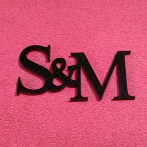 May include: Black wooden letters spelling "S&M" on a pink textured background. The ampersand is between the letters S and M. The letters are in a serif font.