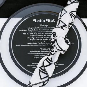 May include: Elegant table setting with black and white plates, silverware, and glassware. A menu with the text "Let's Eat" is visible. Orange flowers and decorative elements complete the sophisticated dining arrangement.