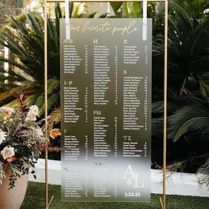 May include: A clear acrylic wedding seating chart with gold lettering and a gold metal frame. The chart lists names alphabetically, with the text "in favorite people" at the top. The date "5.22.22" is at the bottom.