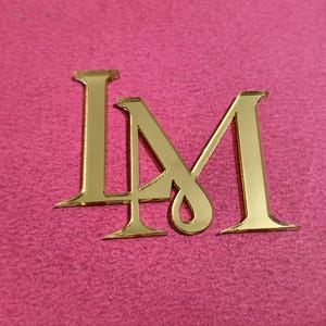 Puede incluir: Letras metálicas doradas 'I' y 'M' sobre un fondo rosa. Las letras están estilizadas y parecen ser un logotipo o monograma. El fondo tiene un aspecto texturizado.