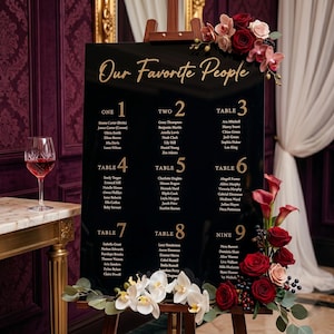 May include: A black acrylic wedding seating chart with gold lettering that reads "Our Favorite People." The chart lists names under table numbers 1-9. Floral arrangements of red and pink roses and orchids decorate the edges.