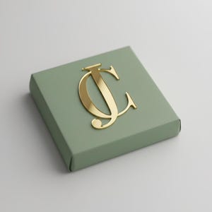 May include: A sage green box with a gold-coloured monogram. The monogram features the letters "J" and "C" in a stylized, elegant font. The box has a simple, clean design, with a matte finish.