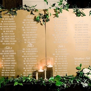 Könnte beinhalten: Goldfarbener Sitzplan mit weißem Text, der die Namen für einen Hochzeitsempfang auflistet. Der Plan ist in Tische von 1 bis 6 unterteilt. Der Plan ist mit Grünpflanzen und weißen Blumen dekoriert.