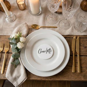 Może przedstawiać: Elegancka aranżacja stołu z białymi talerzami, złotymi sztućcami i kryształowymi kieliszkami. Lniana serwetka ozdobiona jest białymi różami i eukaliptusem. Wizytówka z napisem "Charlotte". Świece i złota łyżka dopełniają aranżację.