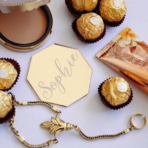 Puede incluir: Arreglo de productos de belleza y bombones. Incluye una polvera, una tarjeta dorada con el nombre "Sophie", un tubo de loción corporal y varios bombones. También hay una pulsera dorada.