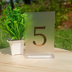 Puede incluir: Un número de mesa de vidrio esmerilado con el número "5" impreso en dorado sobre un fondo degradado verde. El número de mesa está sobre una mesa de madera junto a una planta en maceta.