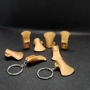 May include: Several gold-coloured, 3D-printed miniature axe-shaped items are displayed. Some are individual pieces, while others are attached to silver keyrings. The axe heads have a textured surface and a detailed design, possibly for a fantasy theme.