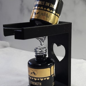 Op de afbeelding: Twee flesjes met heldere nagellak met gouden etiketten. De etiketten hebben de tekst "Magpie" en "Give Me Strength". De flesjes staan in een zwarte hartvormige houder.