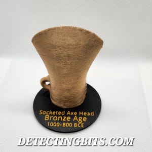 May include: A bronze-coloured socketed axe head replica, with a flared top and a small handle, sits on a black circular base. The base features text in gold lettering: "Socketed Axe Head Bronze Age 1000-800 BCE". The website "DETECTINGBITS.COM" is also visible.