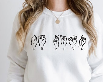 May include: White sweatshirt with the words "BE KIND" spelled out in black American Sign Language. The sweatshirt is a crew neck style and has long sleeves. The model is wearing a gold necklace.