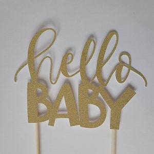 May include: A gold glitter "hello BABY" cake topper. The words are in a cursive font and block letters. Two wooden sticks are attached to the bottom for insertion into a cake or other dessert.