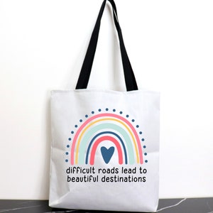 May include: White tote bag with black handles featuring a rainbow design with a blue heart in the center. The text "difficult roads lead to beautiful destinations" is printed below the rainbow.