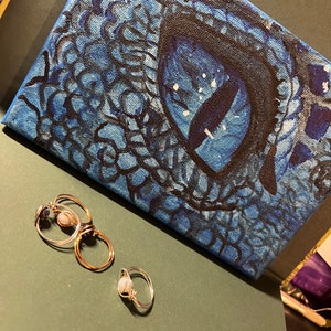 以下が含まれることがあります： 青いキャンバスに黒で描かれた、ドラゴンの目をクローズアップした絵画。手前に宝石がついた金の指輪が3つある。