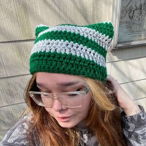May include: A green and white striped crocheted beanie with cat ears. The beanie is made with a soft yarn and has a comfortable fit.