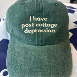 May include: A green baseball cap featuring the embroidered phrase "I have post-cottage depression" in white. The cap has a curved peak and a button on top. The fabric looks like a washed cotton or similar material.