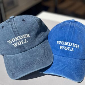 May include: Two blue baseball caps. One is a faded denim blue, the other a bright blue. Both caps feature the white embroidered text "WONDER WOLL" across the front. A casual accessory for everyday wear.