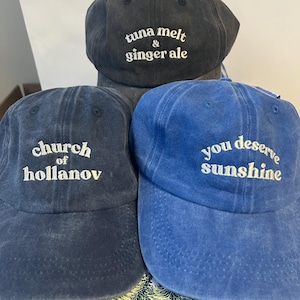 May include: Three baseball caps in shades of blue and gray. The top cap is gray with the text "tuna melt & ginger ale". The middle cap is navy blue with "church of hollanov". The bottom cap is blue with "you deserve sunshine".