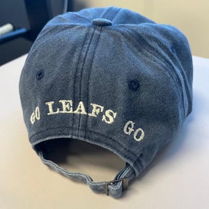 May include: A denim blue baseball cap with the words "GO LEAFS GO" embroidered in white. The cap has a curved brim and an adjustable strap at the back.