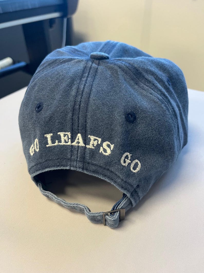 May include: A denim blue baseball cap with the words "GO LEAFS GO" embroidered in white. The cap has a curved brim and an adjustable strap at the back.