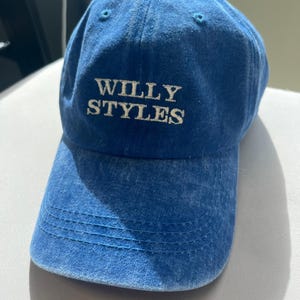 May include: A blue baseball cap with the text "WILLY STYLES" embroidered in white. The cap has a curved brim and a fabric strap closure. The hat is made of a soft, faded denim material.