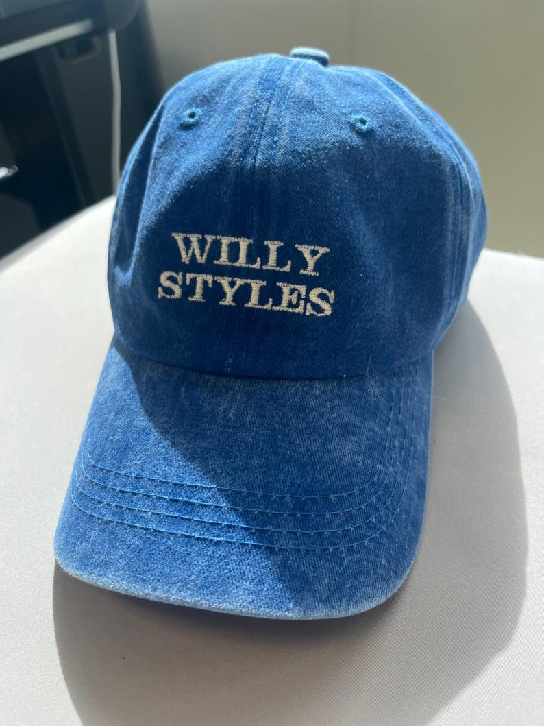 May include: A blue baseball cap with the text "WILLY STYLES" embroidered in white. The cap has a curved brim and a fabric strap closure. The hat is made of a soft, faded denim material.