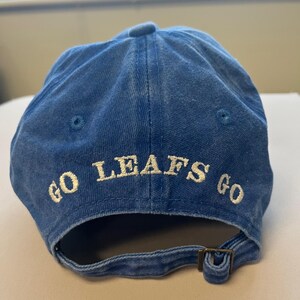 Pu&ograve; includere: Cappellino da baseball blu con le parole "GO LEAFS GO" ricamate in bianco. Il cappellino ha una visiera curva e una chiusura con fibbia metallica regolabile. Realizzato in tessuto morbido e usurato.