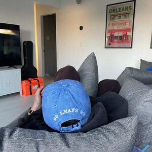 Pu&ograve; includere: Un cappellino da baseball blu con la scritta "GO LEAFS GO" ricamata in bianco. Il cappellino &egrave; indossato al contrario. L'immagine mostra anche un cuscino grigio e una tazza viola con una cannuccia blu.