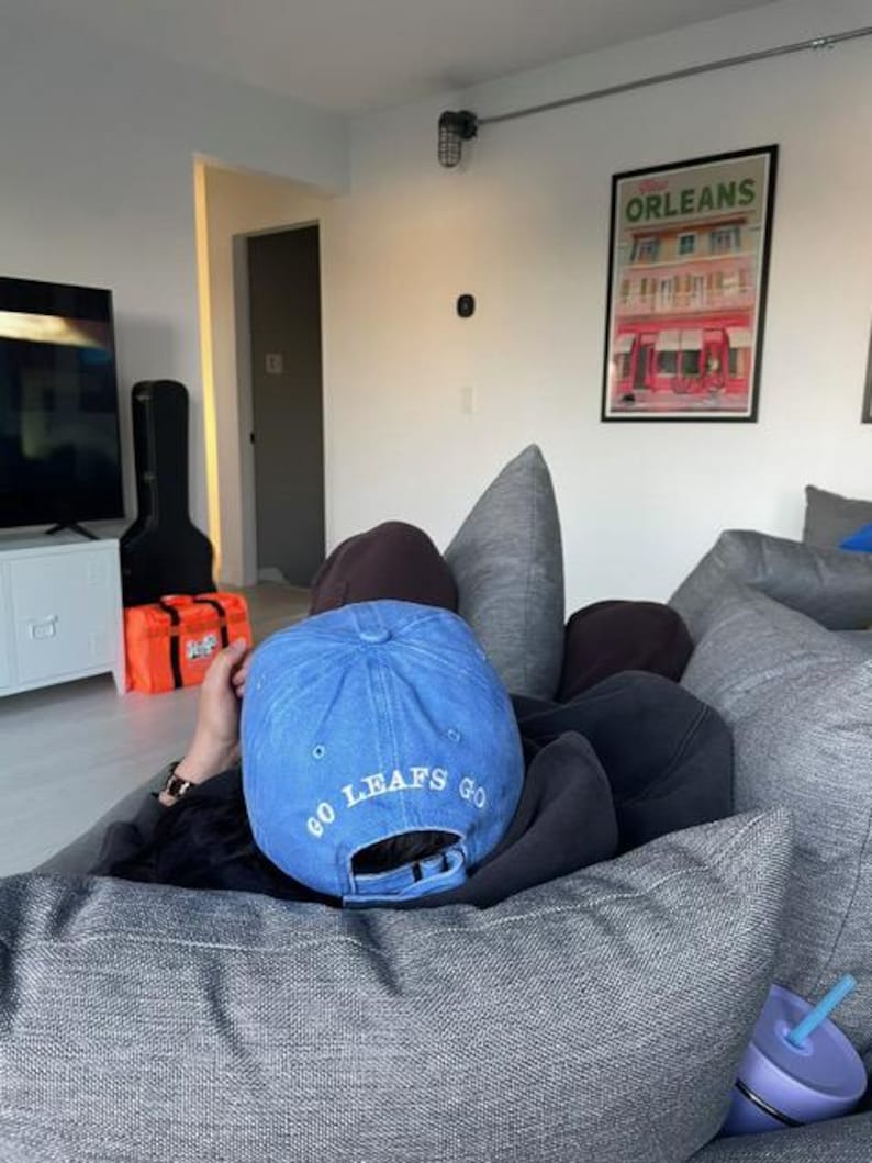 Pu&ograve; includere: Un cappellino da baseball blu con la scritta "GO LEAFS GO" ricamata in bianco. Il cappellino &egrave; indossato al contrario. L'immagine mostra anche un cuscino grigio e una tazza viola con una cannuccia blu.