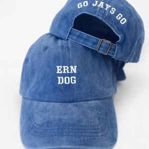 May include: Two blue baseball caps, one on top of the other. The front cap displays "ERN DOG" in white, while the top cap reads "GO JAYS GO". Both caps feature a curved peak and an adjustable strap with a metal buckle.