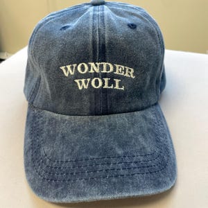 Pu&ograve; includere: Cappellino da baseball in denim blu con la scritta "WONDER WOLL" ricamata in bianco. Il cappellino ha una visiera curva e un bottone sulla parte superiore. Un cappello casual.