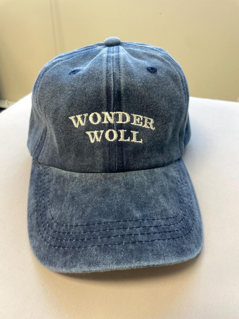 Pu&ograve; includere: Cappellino da baseball in denim blu con la scritta "WONDER WOLL" ricamata in bianco. Il cappellino ha una visiera curva e un bottone sulla parte superiore. Un cappello casual.