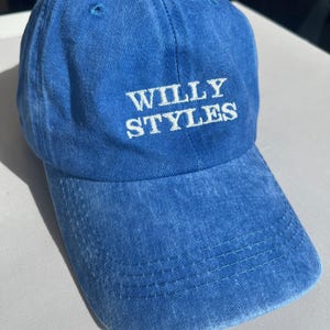 May include: A blue baseball cap with the text "WILLY STYLES" embroidered in white. The cap has a curved brim and a small button on top. The fabric appears to be a soft, washed cotton.