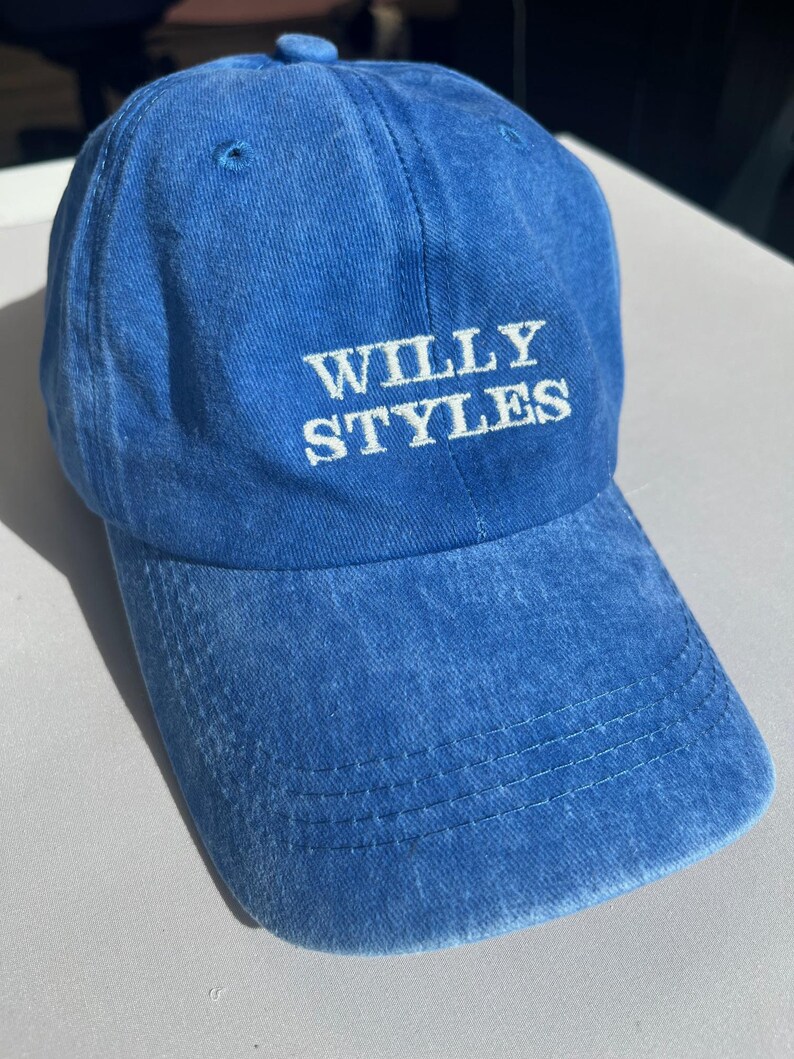 May include: A blue baseball cap with the text "WILLY STYLES" embroidered in white. The cap has a curved brim and a small button on top. The fabric appears to be a soft, washed cotton.
