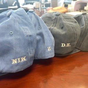 Pu&ograve; includere: Quattro cappellini da baseball in tonalit&agrave; blu e grigie sono esposti su una superficie di legno. Ogni cappellino ha una visiera curva e un bottone sulla parte superiore. I cappellini sono ricamati con testo bianco: "NIK", "Z", "D.H." e "S.T."