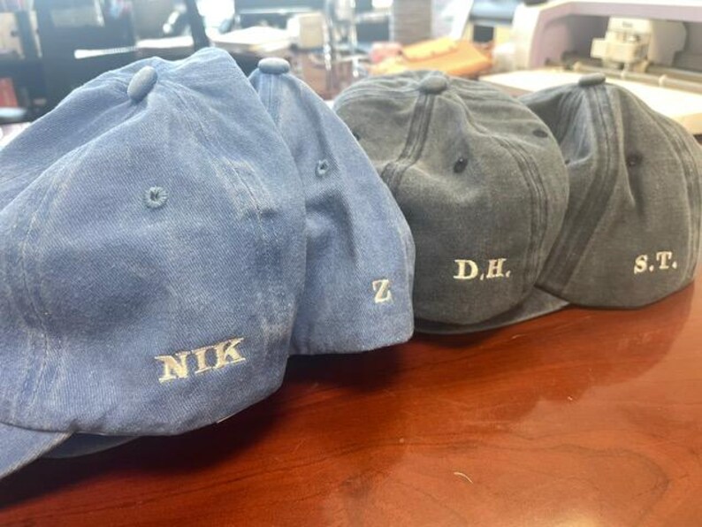 Pu&ograve; includere: Quattro cappellini da baseball in tonalit&agrave; blu e grigie sono esposti su una superficie di legno. Ogni cappellino ha una visiera curva e un bottone sulla parte superiore. I cappellini sono ricamati con testo bianco: "NIK", "Z", "D.H." e "S.T."