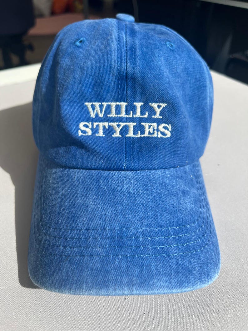 May include: A blue baseball cap with the text "WILLY STYLES" embroidered in white. The cap has a curved brim and a slightly faded appearance, suggesting a vintage or worn look. The cap is made of a soft, durable fabric.