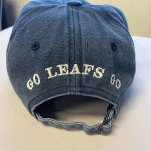May include: A denim blue baseball cap with the words "GO LEAFS GO" embroidered in white across the back. The cap has a curved brim and an adjustable strap.
